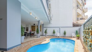 A swimming pool with a curved edge, surrounded by a tiled deck. There are potted plants and a white chair near the pool. - Square One Hotel Patong (Patong)