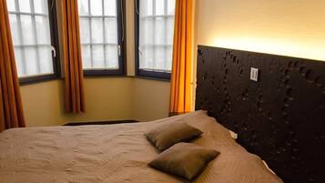 A bed with a textured beige cover and two pillows, a dark headboard with a textured pattern, and a window with orange curtains.