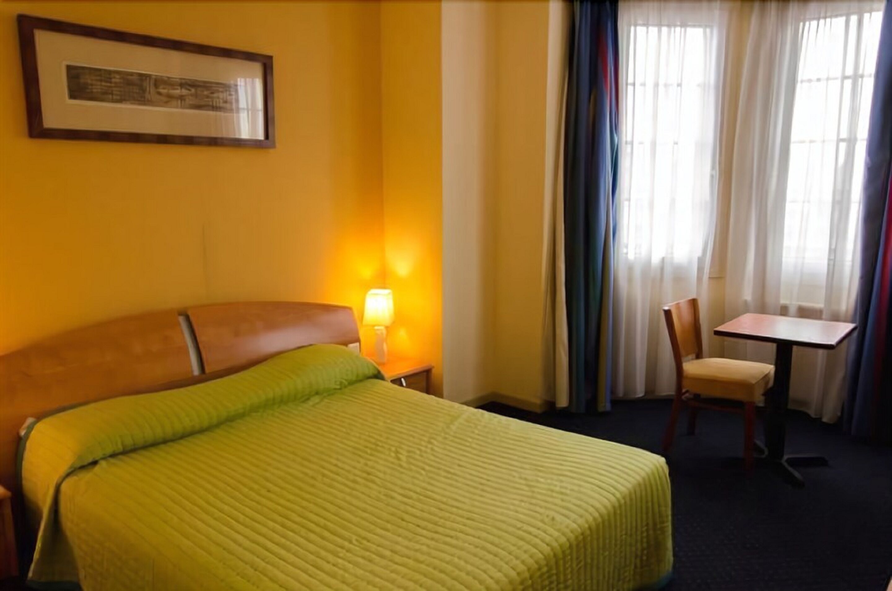 A hotel room with a bed, a nightstand with a lamp, a chair, a table, and a window with curtains.