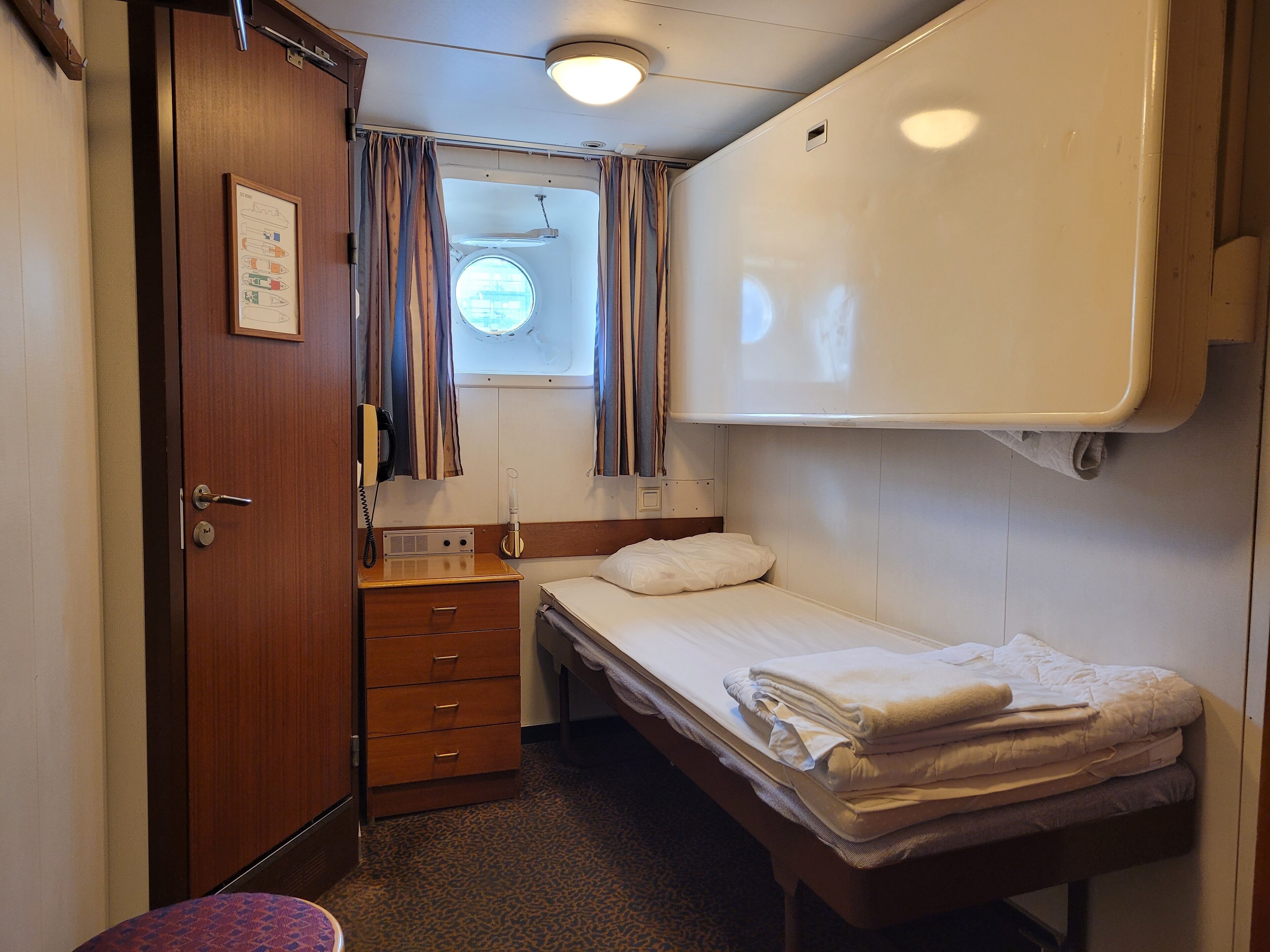 Un dormitorio compacto con una cama individual, una mesita de noche pequeña, una cómoda con cajones, una ventana con cortinas y una lámpara en el techo.