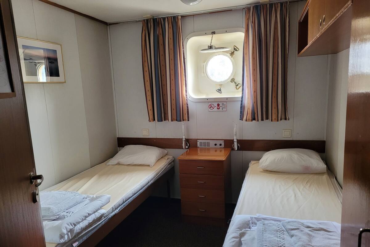 Una cabaña compacta con dos camas individuales, una ventana pequeña con cortinas, un aparador de madera y un cuadro enmarcado en la pared.