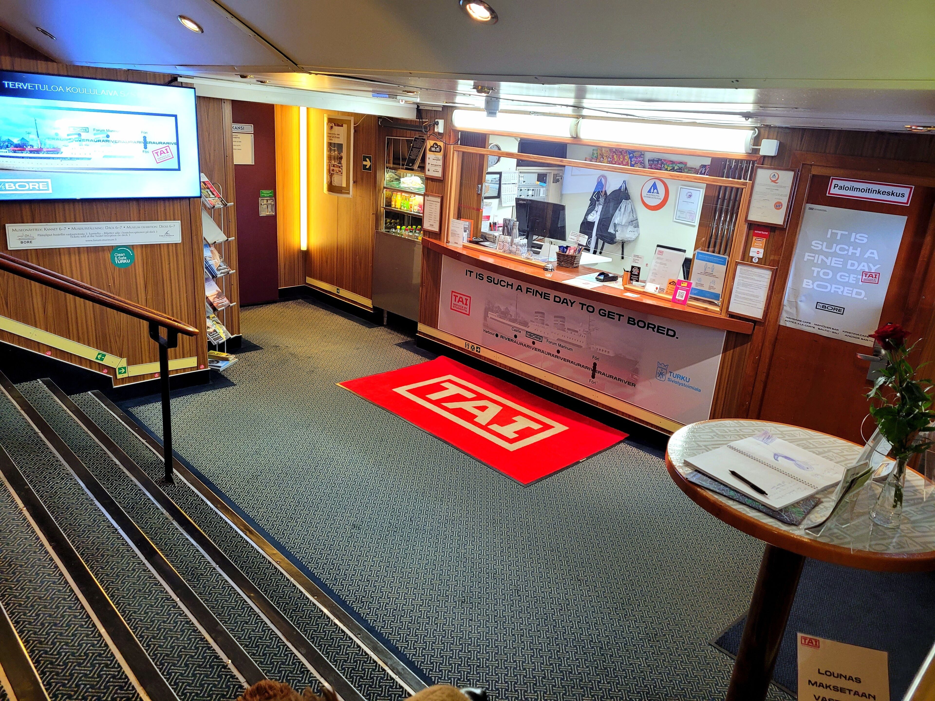 A hotel lobby with a reception desk, a digital screen displaying information, a red carpet with a logo, and a staircase leading to other areas.