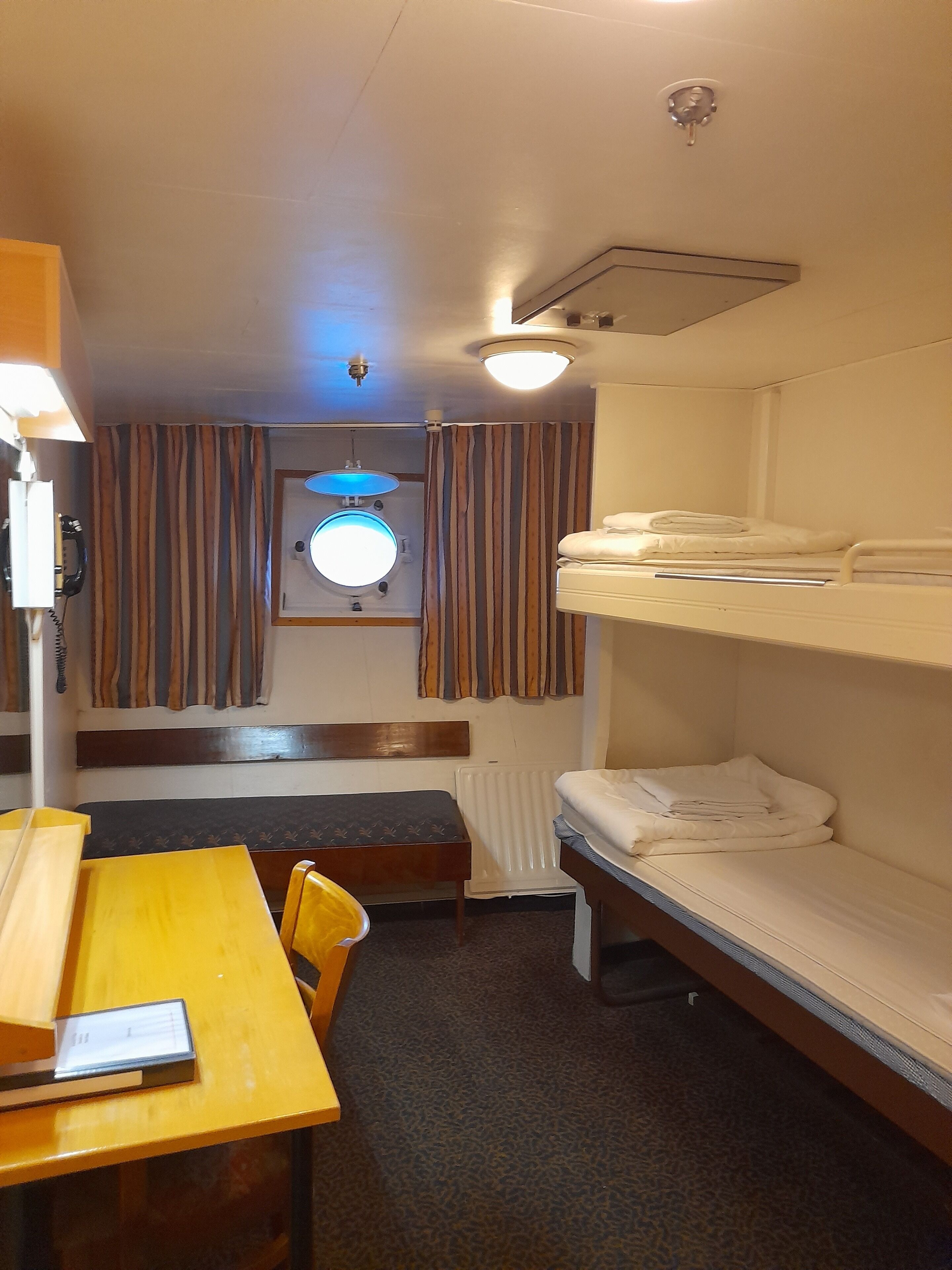 A compact hotel room with a bunk bed, a small table with a chair, a desk, and a window with curtains.