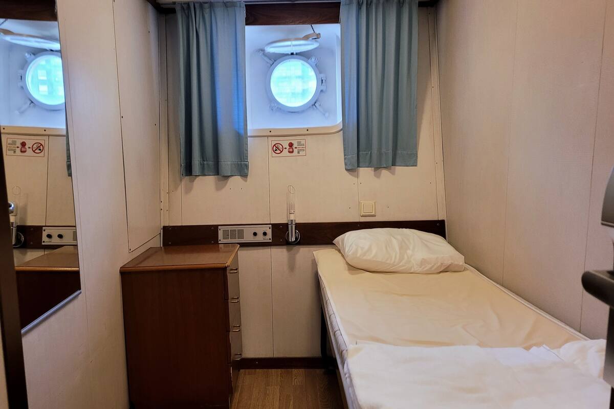 Una habitación pequeña y funcional con una cama individual, un escritorio de madera, un armario pequeño y una ventana con cortinas.