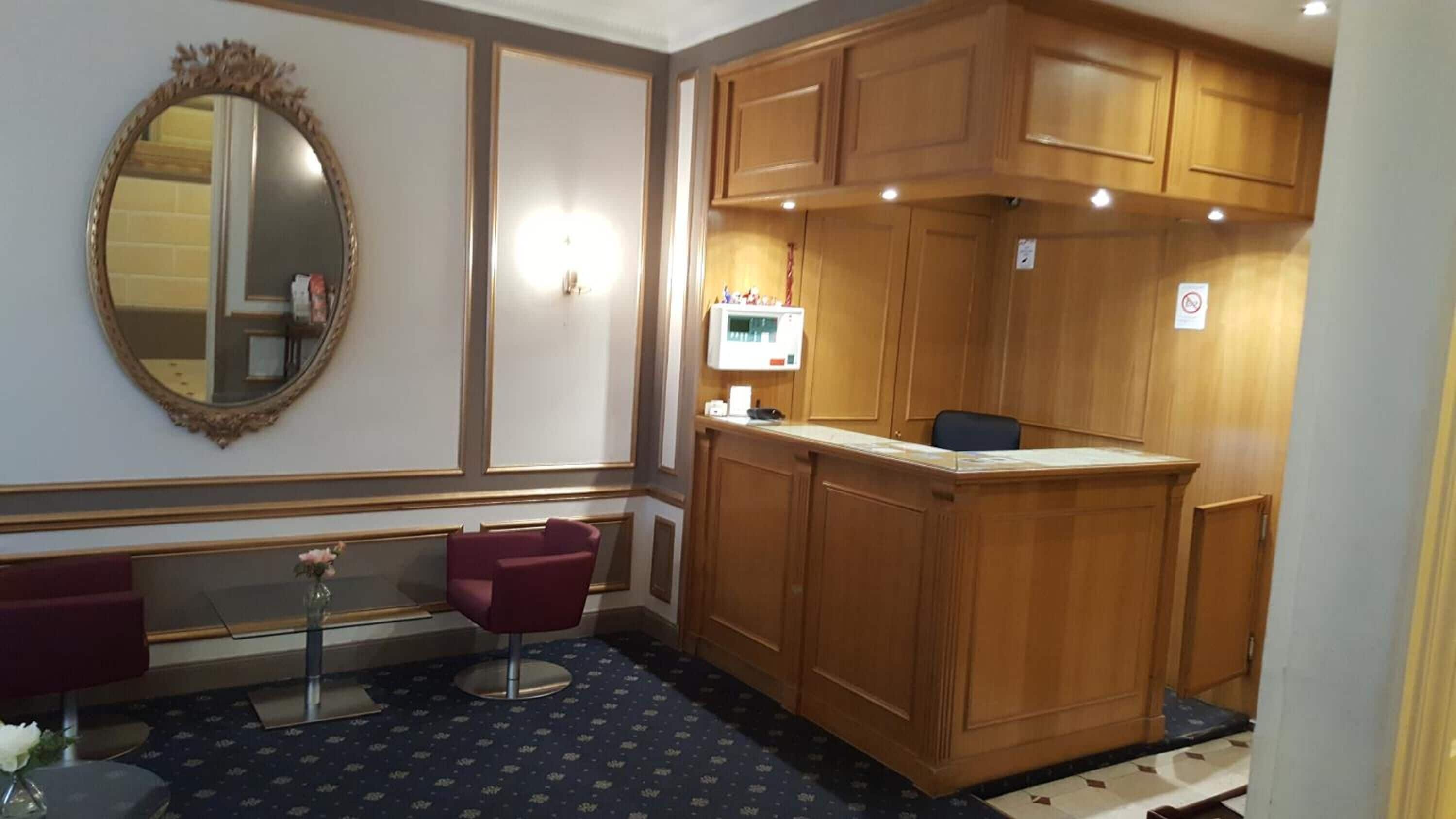 A hotel room with a wooden vanity, a mirror, a small table with a vase, and a patterned carpet.