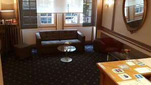Een kamer met een bank, een kleintje tafeltje met een vaas, een rode stoel, een houten bureau met papieren, en een grote spiegel.