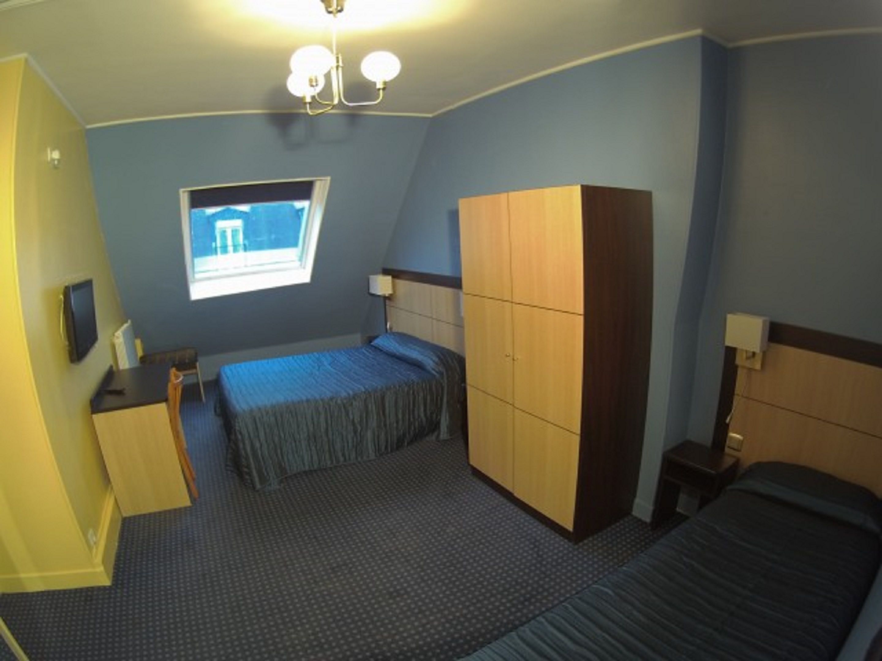 A small, well-lit bedroom with two beds, a wardrobe, a desk, and a window providing a view.