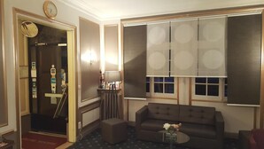 Hotelkamer met een bank, een bijzettafeltje met bloemen, een lamp, een groot raam met ronde patronen, en een deur met een spiegel en een klok.