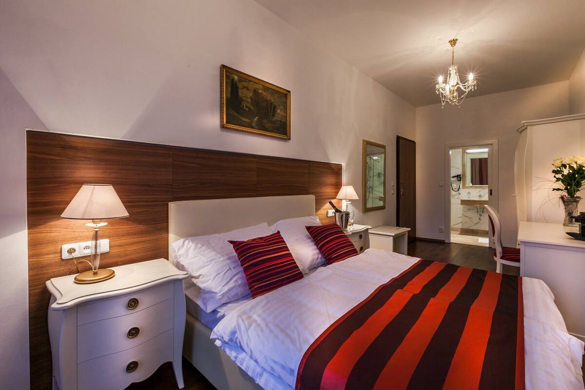 A hotel room with a large bed, two bedside tables with lamps, a wooden headboard, a painting on the wall, and a chandelier.