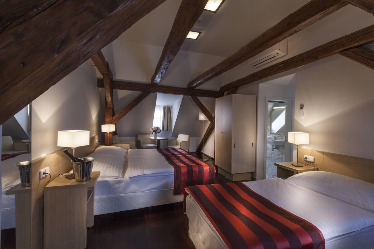 A hotel room with two beds, wooden beams, a small table with a lamp, and a bathroom visible through an open door.
