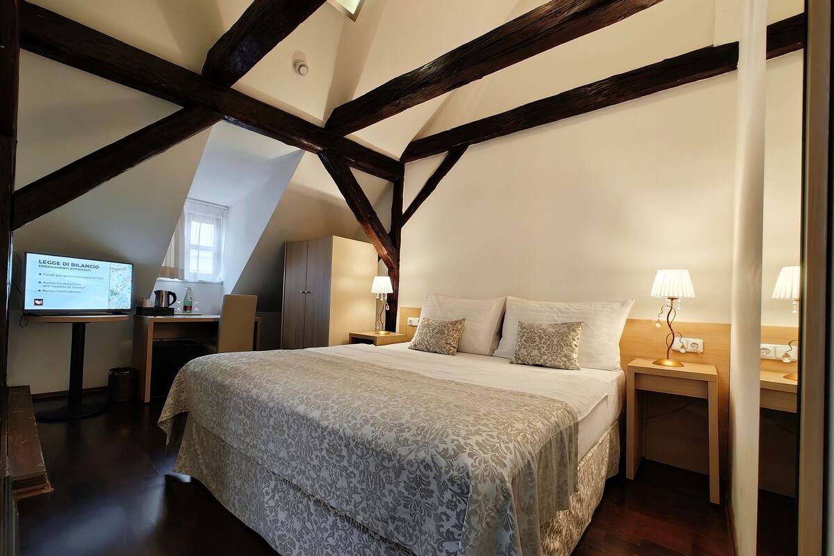 A bedroom with a large bed, wooden beams, a desk with a computer, and a small table with a lamp.