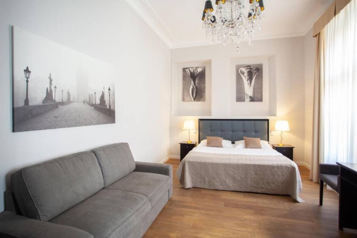 A hotel room with a large bed, a grey sofa, two bedside tables with lamps, and a chandelier.