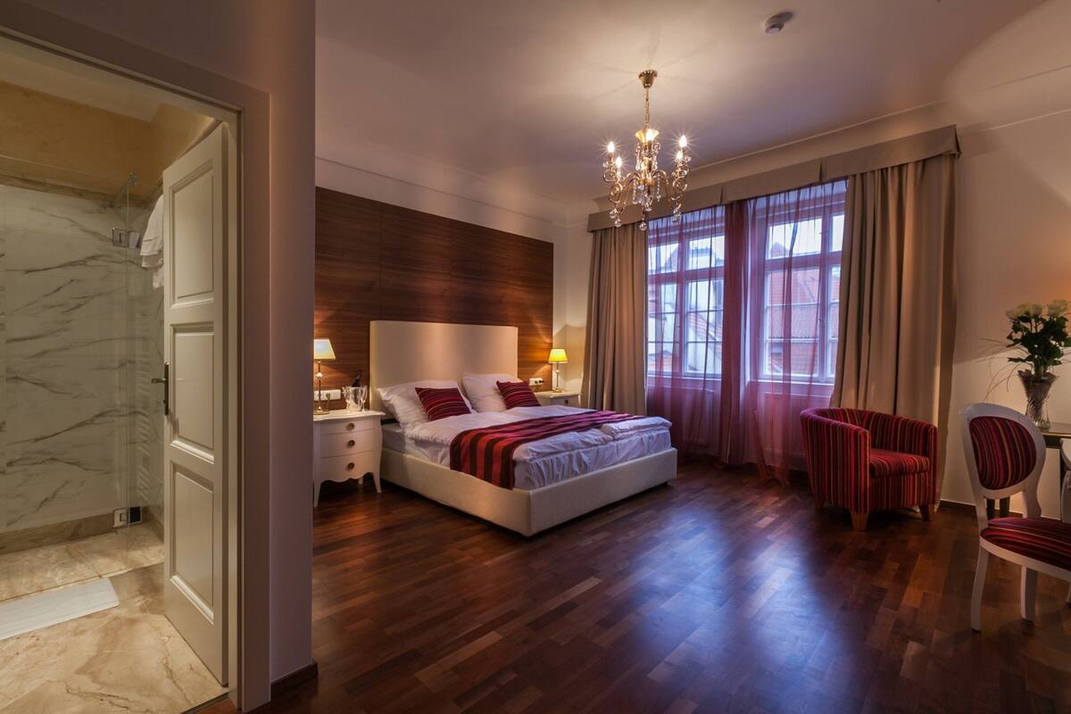 A bedroom with a large bed, wooden panel wall, chandelier, and a view of the outside through large windows.