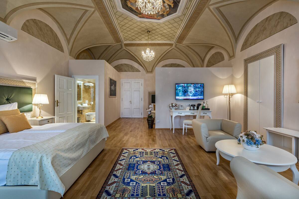 A hotel room with a large bed, a desk, a chair, a TV, and a chandelier.