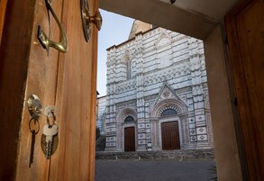 Una vista attraverso un portale in legno con maniglia e chiavi in ottone, che conduce a un edificio con architettura in pietra finemente decorata e due grandi porte in legno.