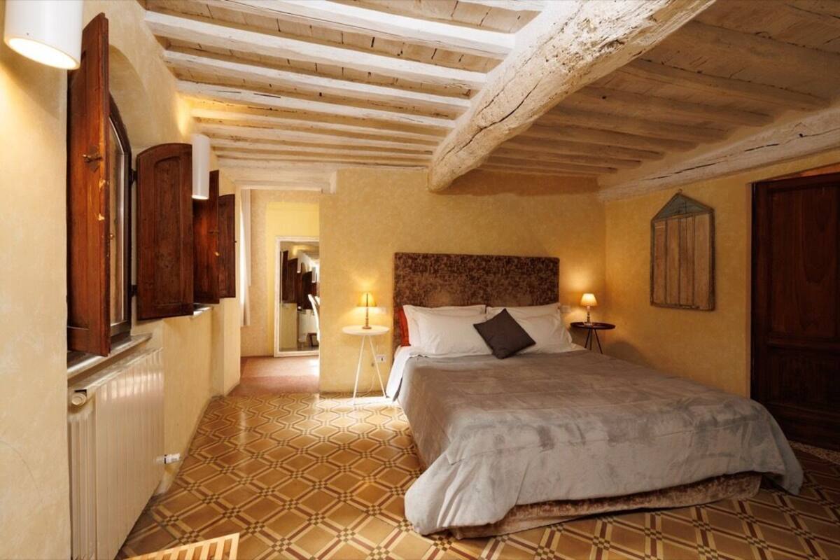 A bedroom with a bed, wooden ceiling beams, patterned floor, and a nightstand with a lamp.