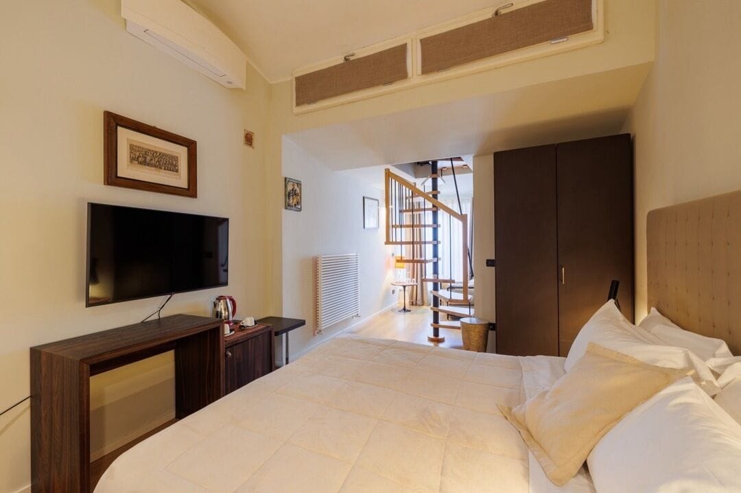 A bedroom with a bed, a TV, a wooden desk, a wardrobe, and a staircase leading to another room.