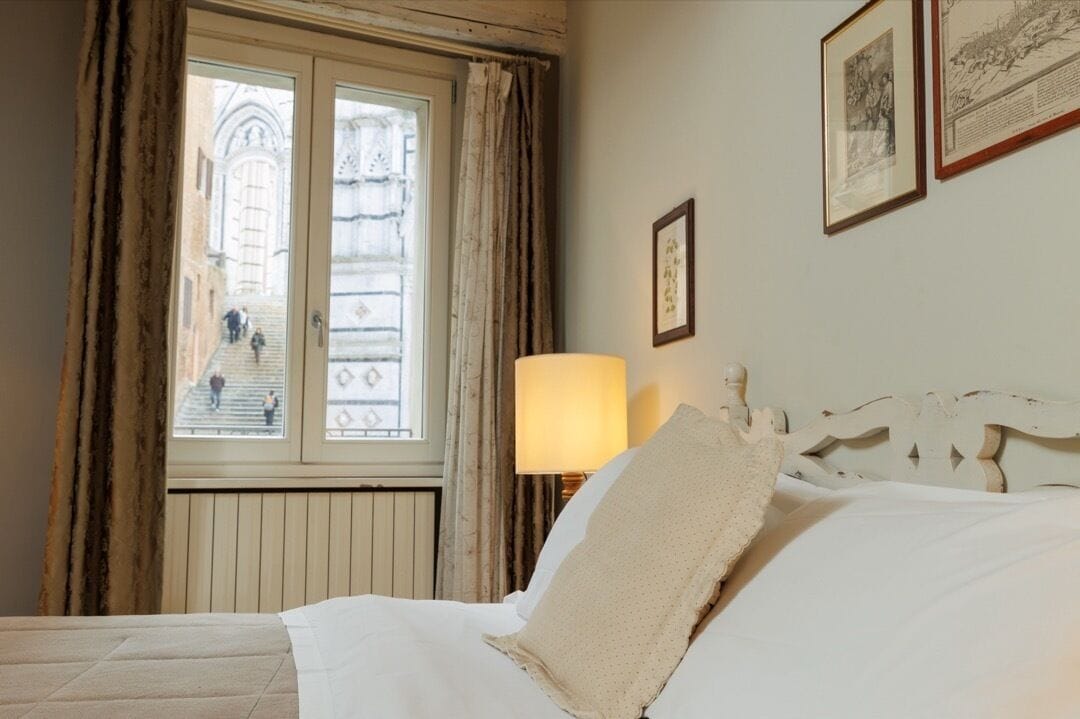 Une chambre d’hôtel avec un lit, une fenêtre avec des rideaux, une lampe et des tableaux encadrés au mur.