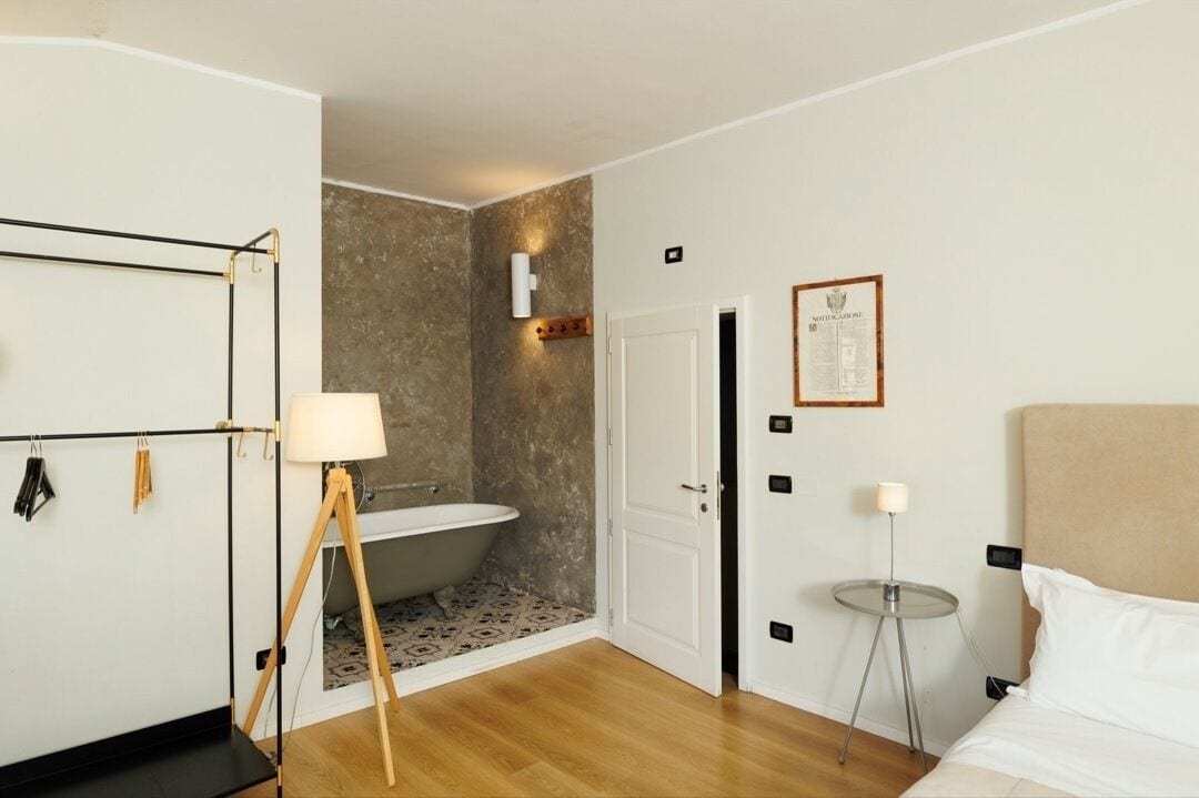 Ein Hotelzimmer mit Holzboden, einem weißen Bett, einem kleinen runden Tisch mit Lampe, einer Wandleuchte, einem gerahmten Zertifikat an der Wand und einer Badewanne in der Ecke.