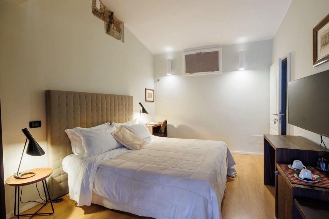 Quarto com cama, mesa de cabeceira, luminária, TV e pequena mesa de madeira.