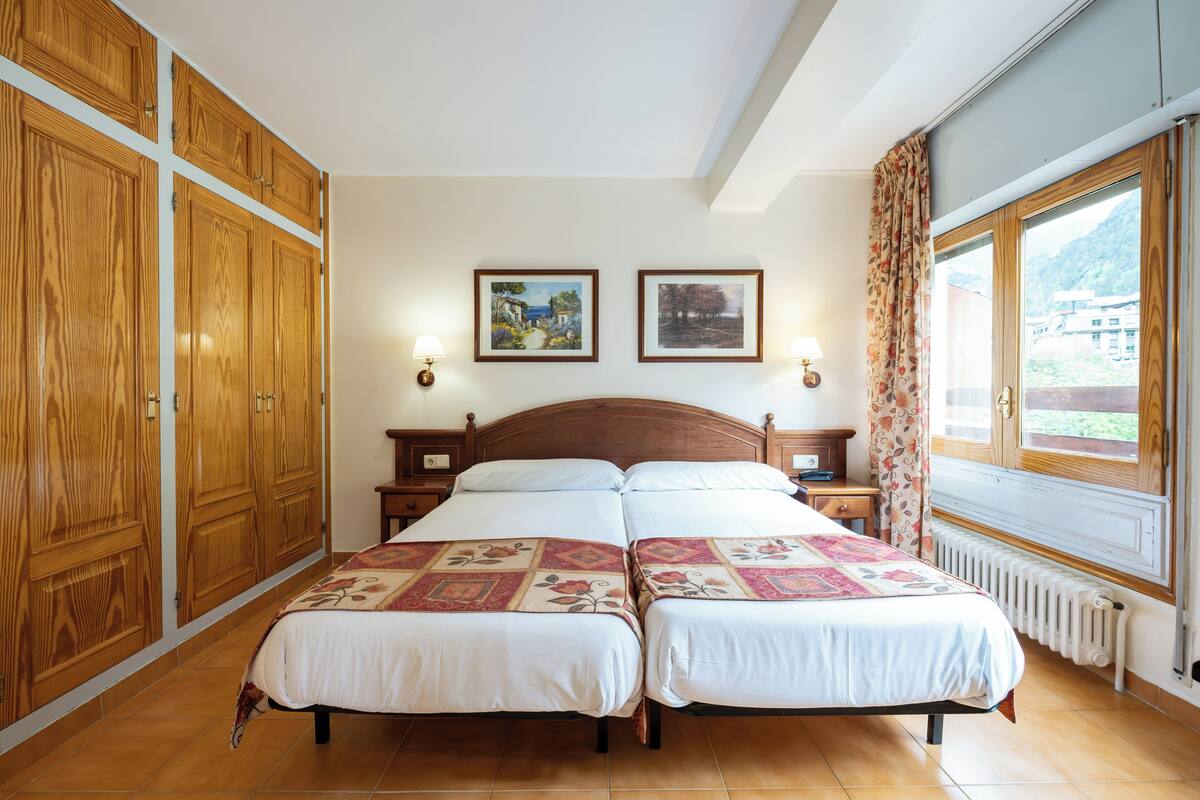 Quarto com duas camas, armários de madeira, cabeceira de madeira, duas imagens emolduradas, janela com cortinas e um radiador.