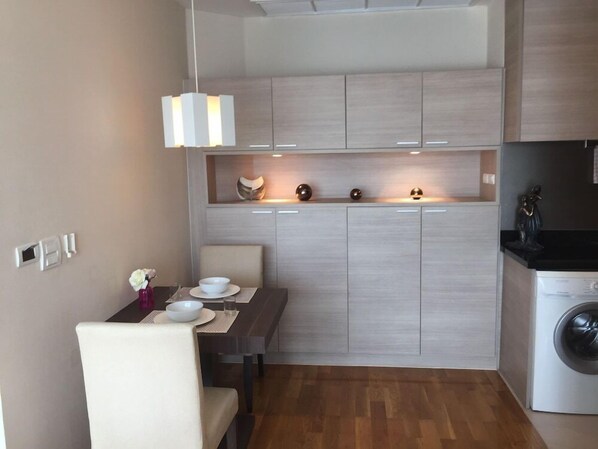 A compact kitchen with a dining area, wooden cabinets, a washing machine, and a pendant light. - 39 Boulevard Executive Residence (Bangkok)