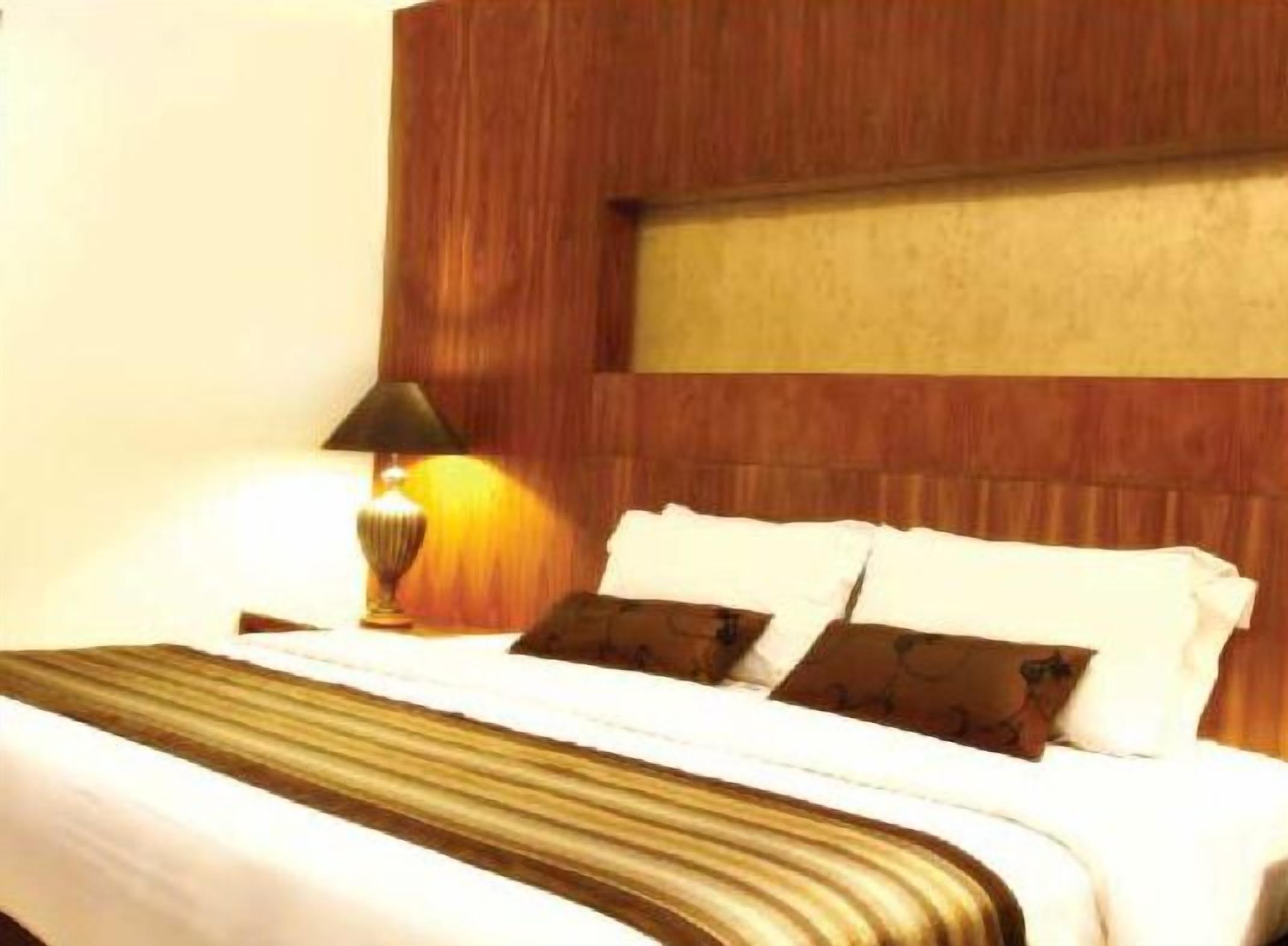 A hotel room with a wooden headboard, a bed with white bedding and brown pillows, and a lamp on a bedside table.