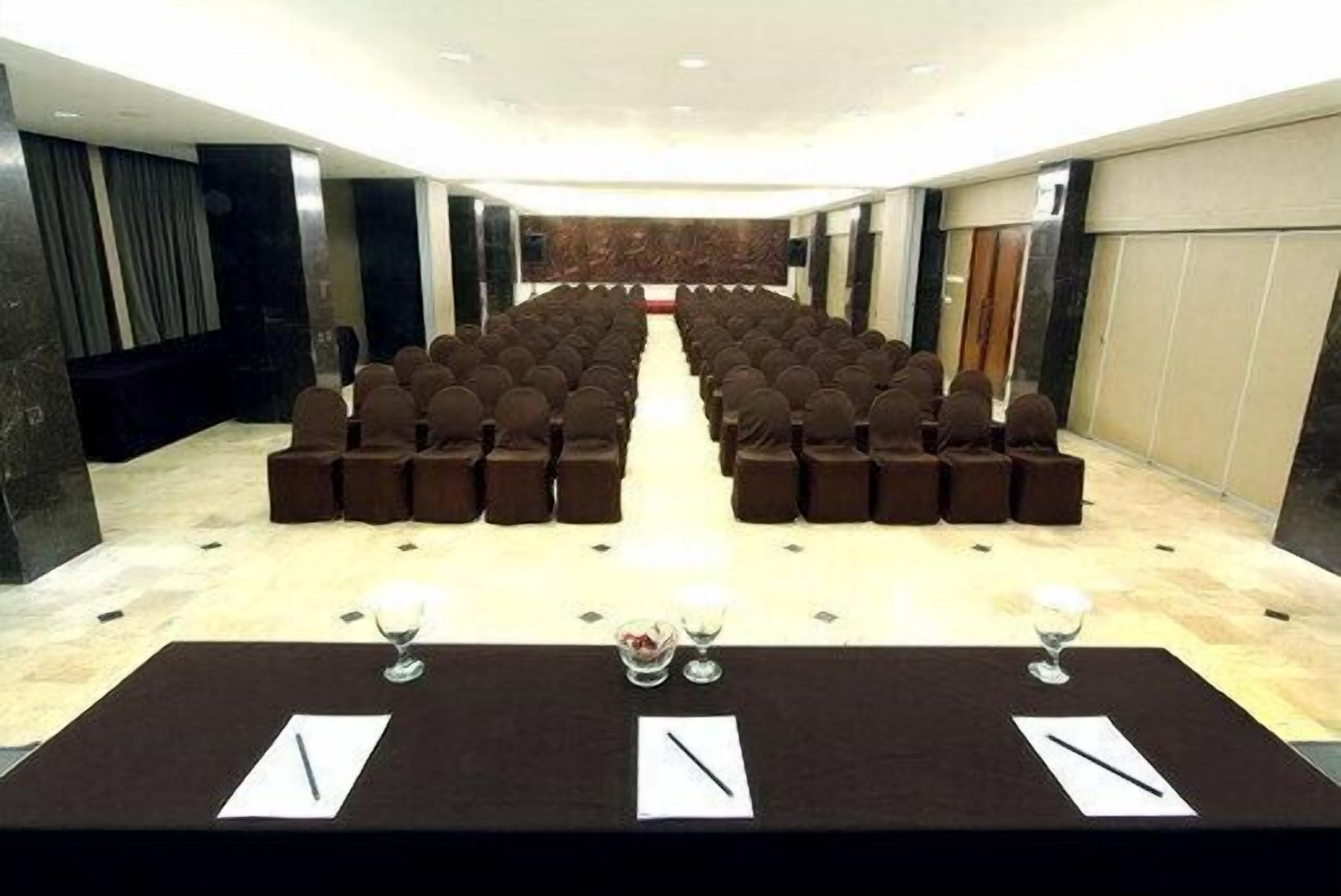 Una sala de conferencias con filas de sillas, una larga mesa con papeles y bolígrafos, y un escenario al frente.