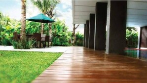 A wooden pathway leading to a covered area with a table and chairs under a green umbrella, surrounded by lush greenery.