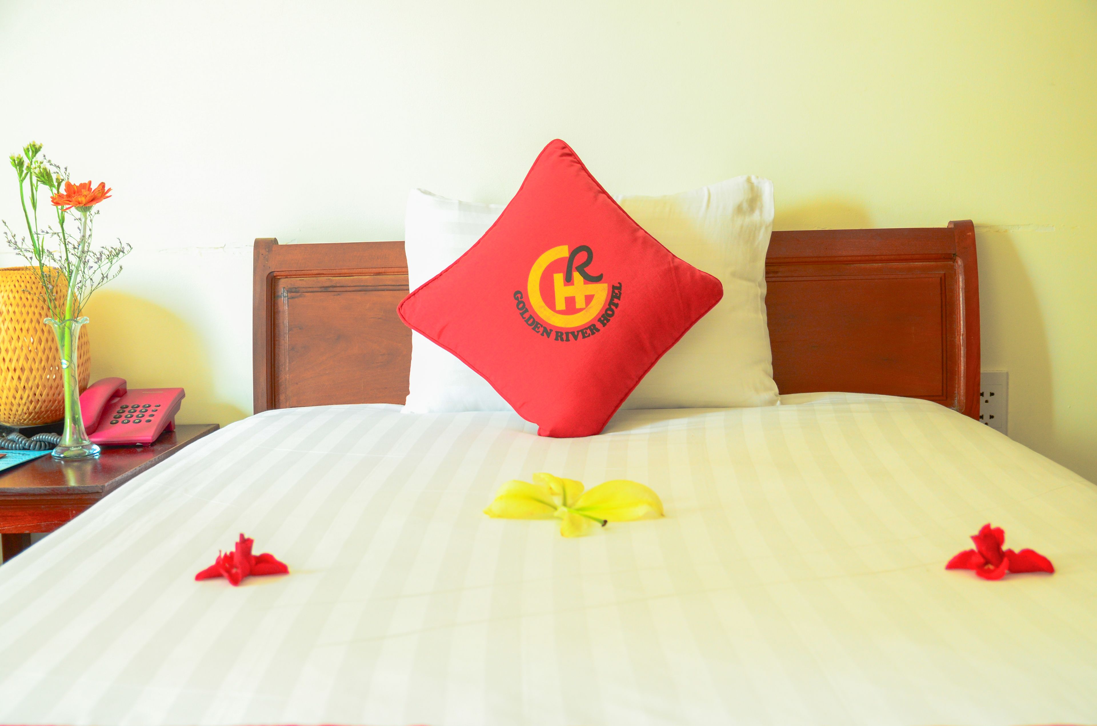 A neatly made bed with a red pillow featuring a logo, a yellow flower, and red rose petals.