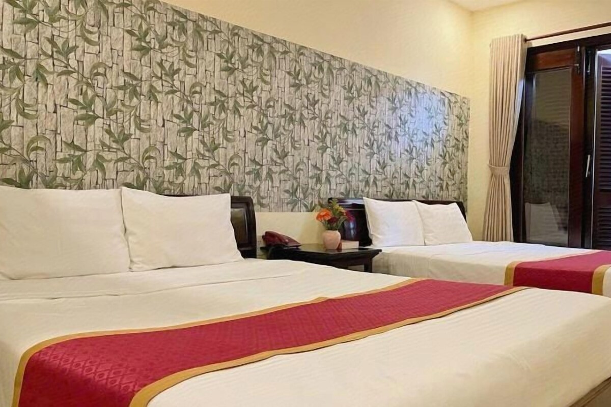 A hotel room with two beds, a patterned wallpaper, a nightstand with a phone, and a window with a view.
