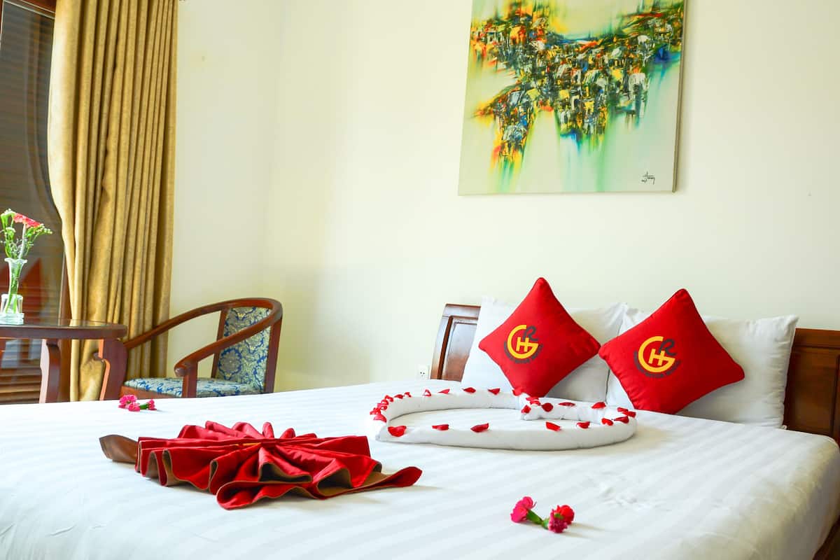 Una cama bien hecha con almohadas rojas y blancas, un lazo rojo y pétalos de rosa. Hay un cuadro en la pared y una silla en la esquina.