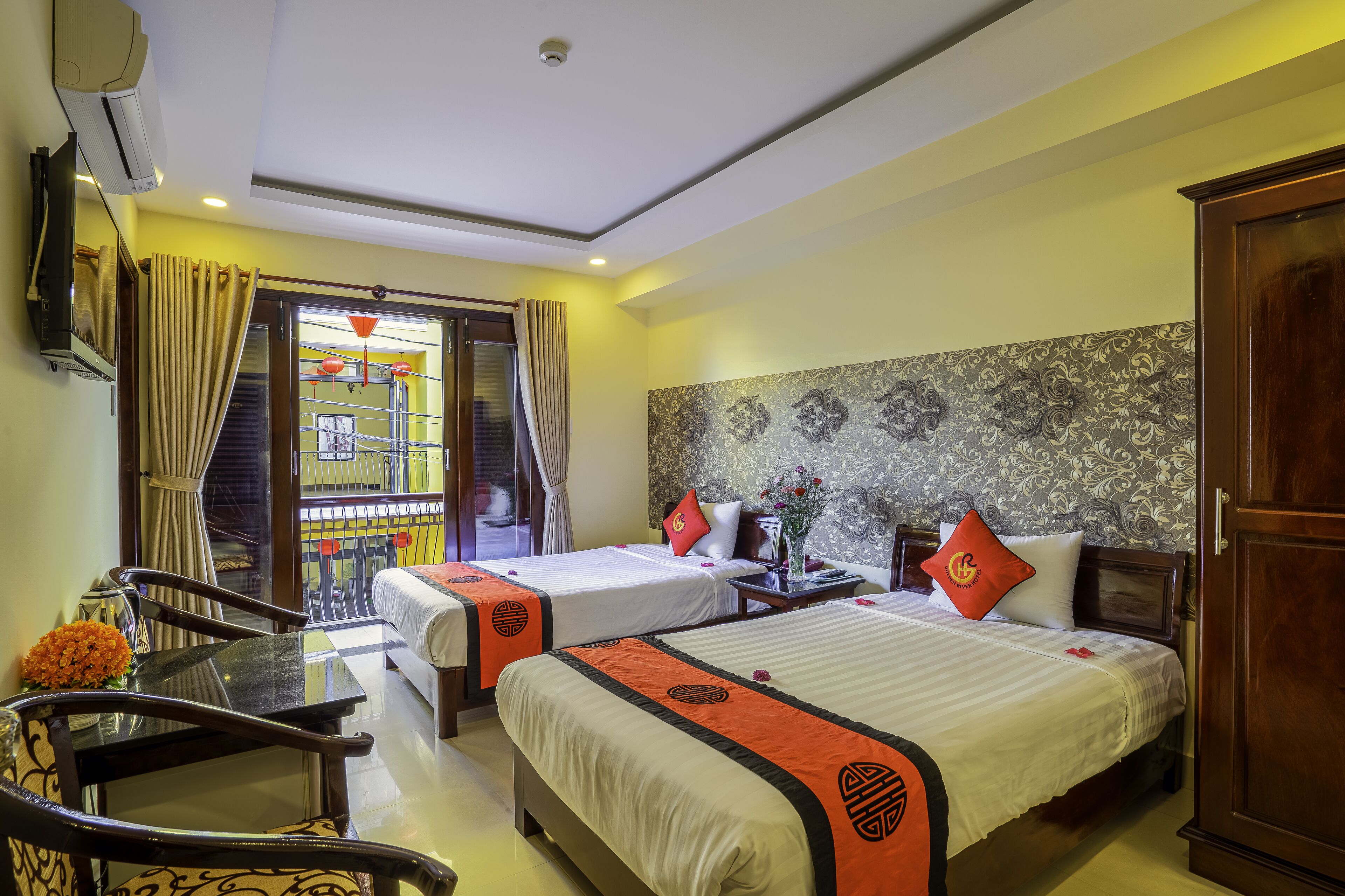 A hotel room with two beds, a dining table, chairs, a TV, and a balcony with a view.