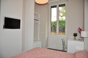 A bedroom with a bed, a TV mounted on the wall, a wardrobe, a desk with a lamp, and a window with a view of greenery. - Dea Roma Inn (Rome)