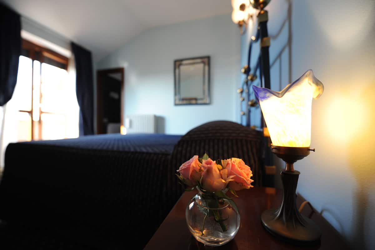 Una habitación de hotel con cama, una mesita de noche con lámpara y un florero, y una ventana con cortinas.