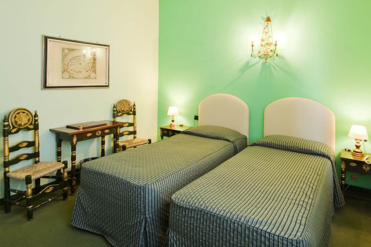 Una habitación con dos camas individuales, una silla de madera, una mesita y una lámpara de pared.