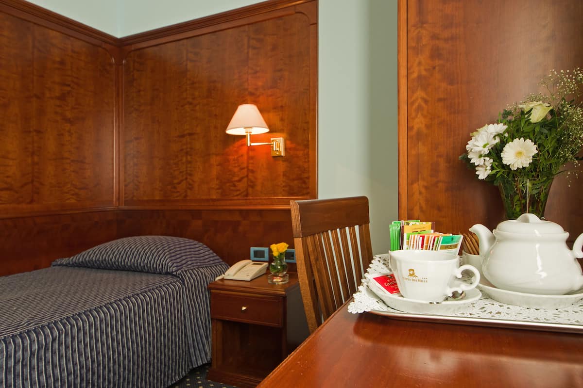 Habitación de hotel con cabecera de madera, mesita de noche con lámpara, teléfono y bandeja con tetera, tazas y un florero con flores.