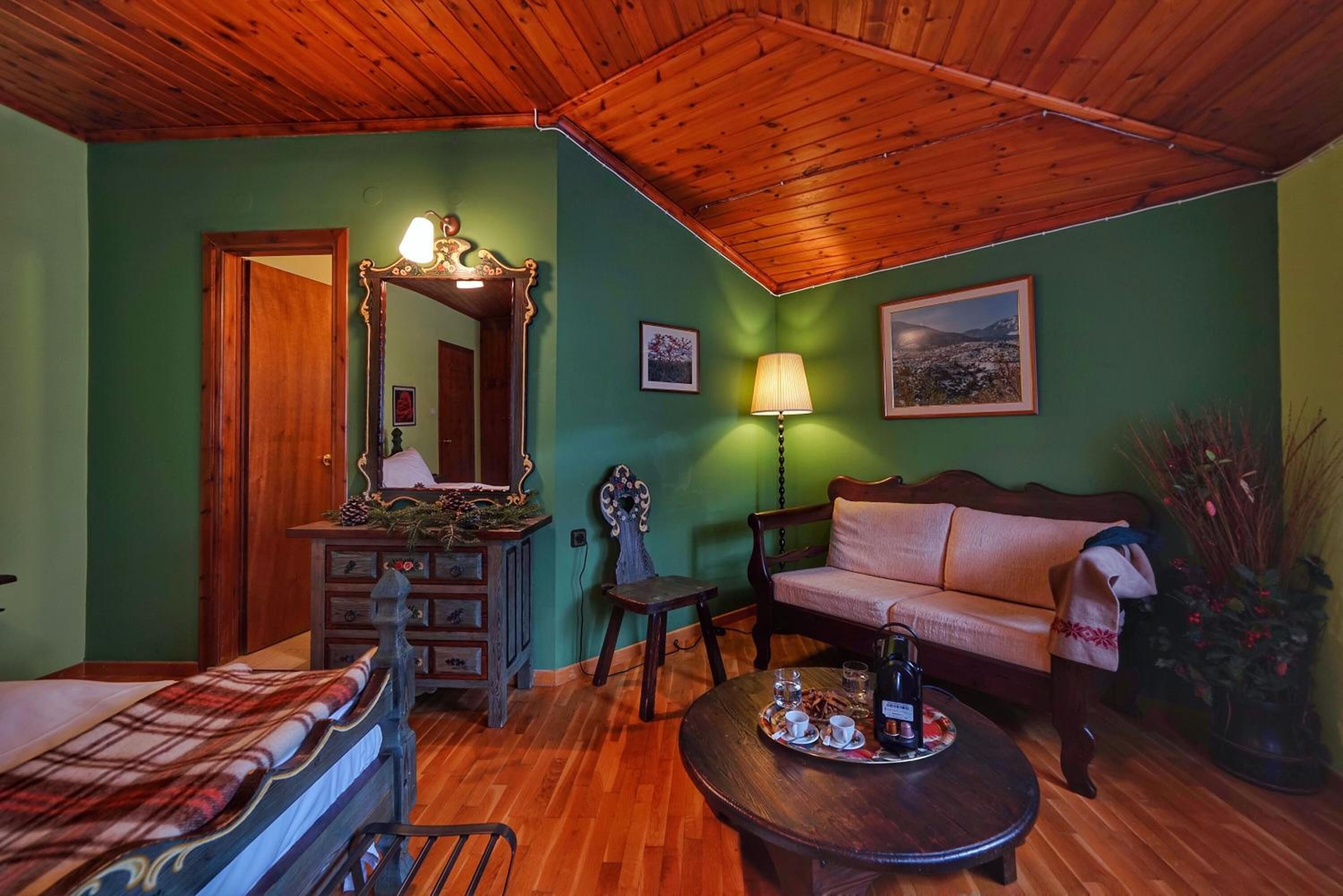 A cozy room with a wooden ceiling, a green wall, a wooden dresser, a sofa, a bed, and a coffee table with a tray of items.