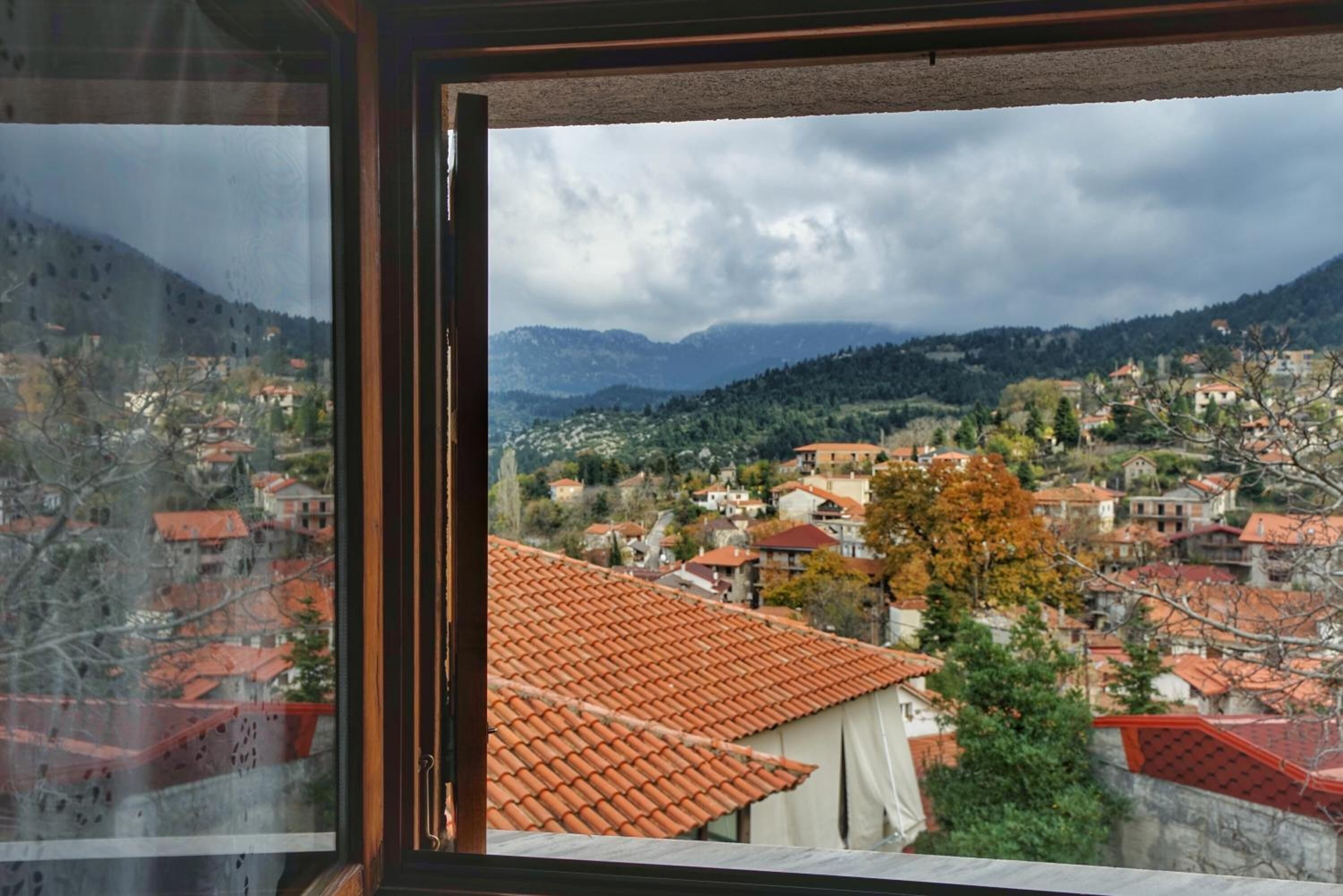 Vista a través de una ventana de un pueblo con tejados rojos, rodeado de colinas y un cielo nublado.