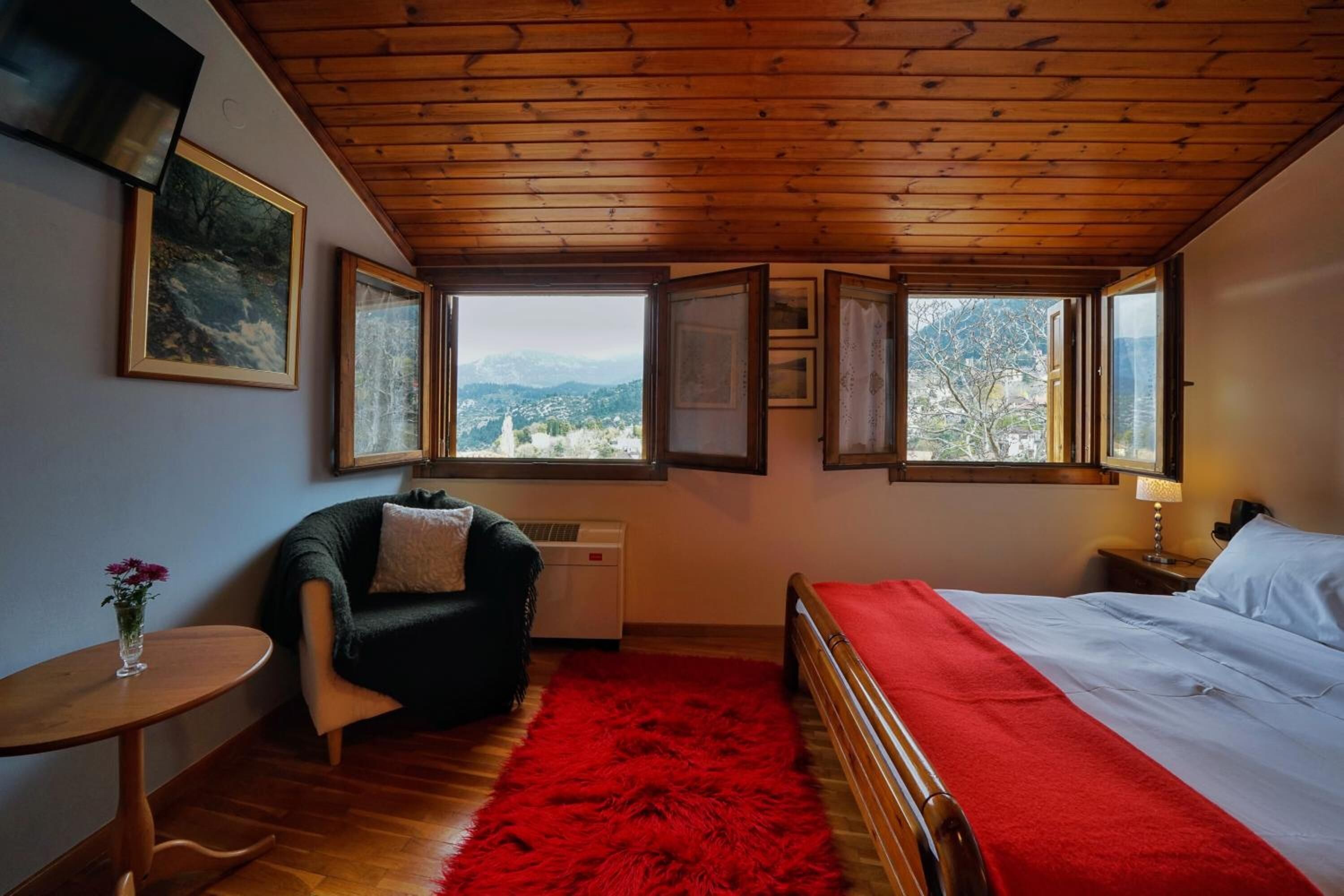 Ein Schlafzimmer mit Holzbalkendecke, einem Bett mit roter Decke, einem Sessel, einem kleinen Tisch mit einer Vase und mehreren Fenstern mit Blick auf die Berge.