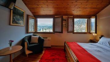 Ein Schlafzimmer mit Holzbalkendecke, einem Bett mit roter Decke, einem Sessel, einem kleinen Tisch mit einer Vase und mehreren Fenstern mit Blick auf die Berge.