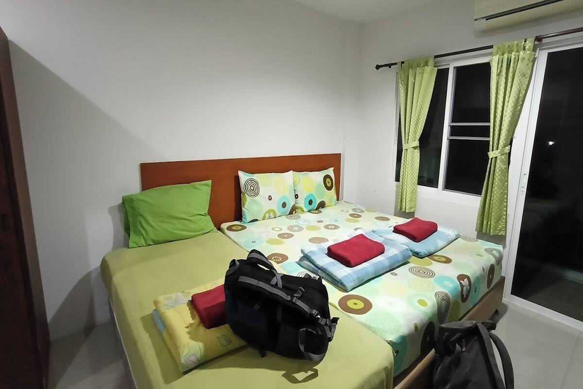 A single bed with a wooden headboard, green and red pillows, a patterned bedspread, and a black backpack on the bed.