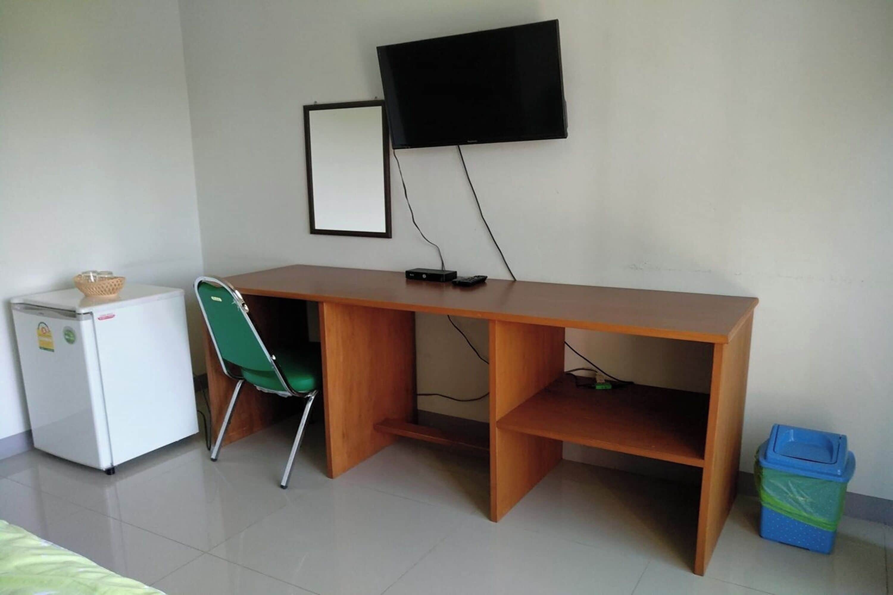 A room with a wooden desk, a green chair, a white refrigerator, a TV mounted on the wall, and a blue trash bin.