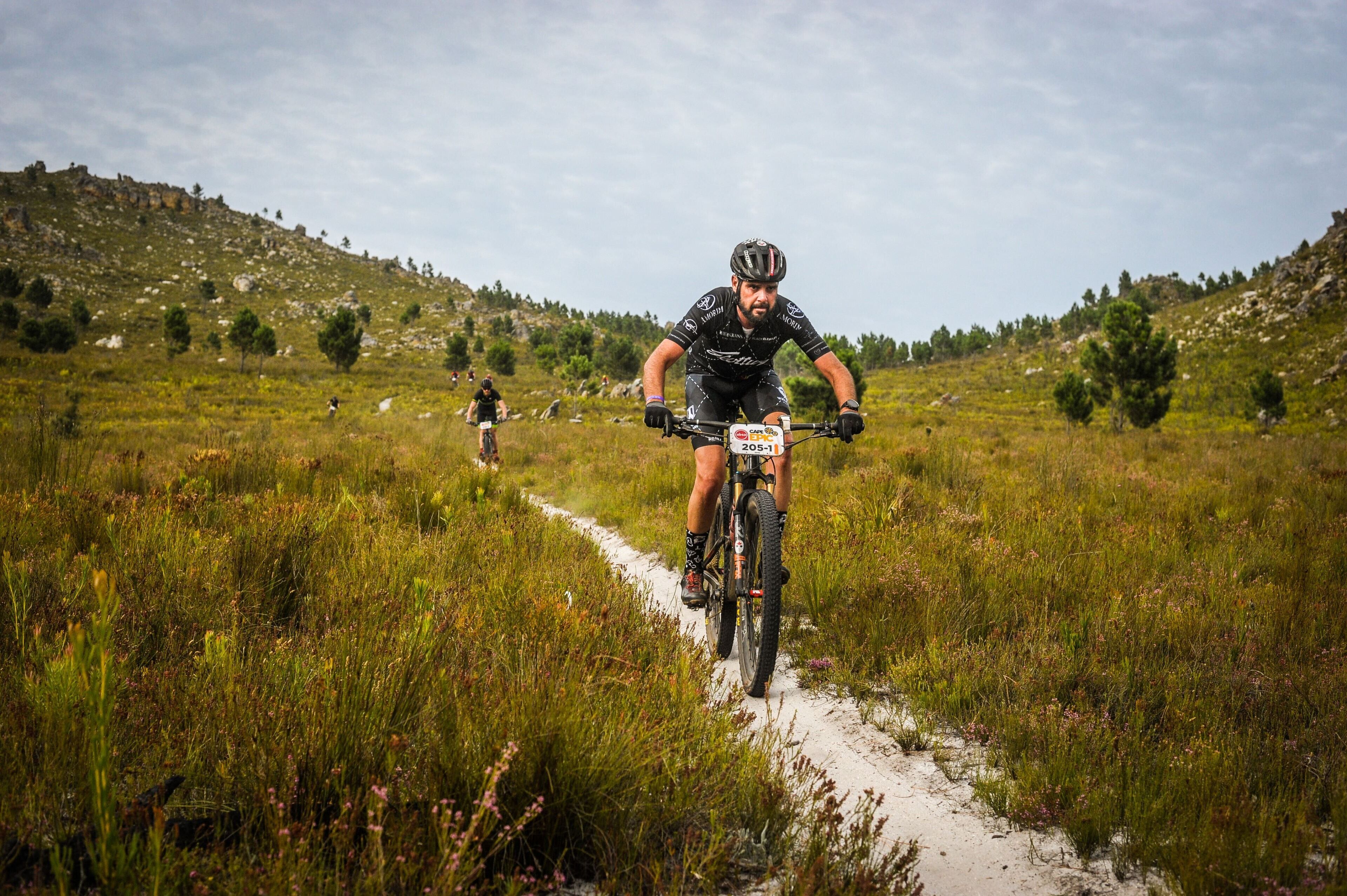 A mountain biker on a dirt trail with a number tag, surrounded by grassy terrain and trees.