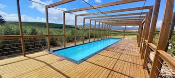 Um deck de madeira com piscina, ladeado por uma pérgula de madeira, com vista para um vinhedo.