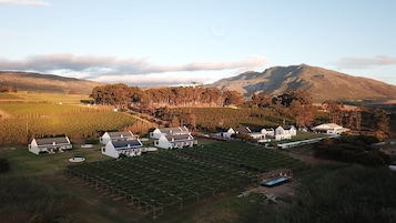 Um complexo vinícola com vários edifícios brancos, incluindo uma casa grande e algumas construções menores, cercado por vegetação exuberante e um cenário montanhoso.