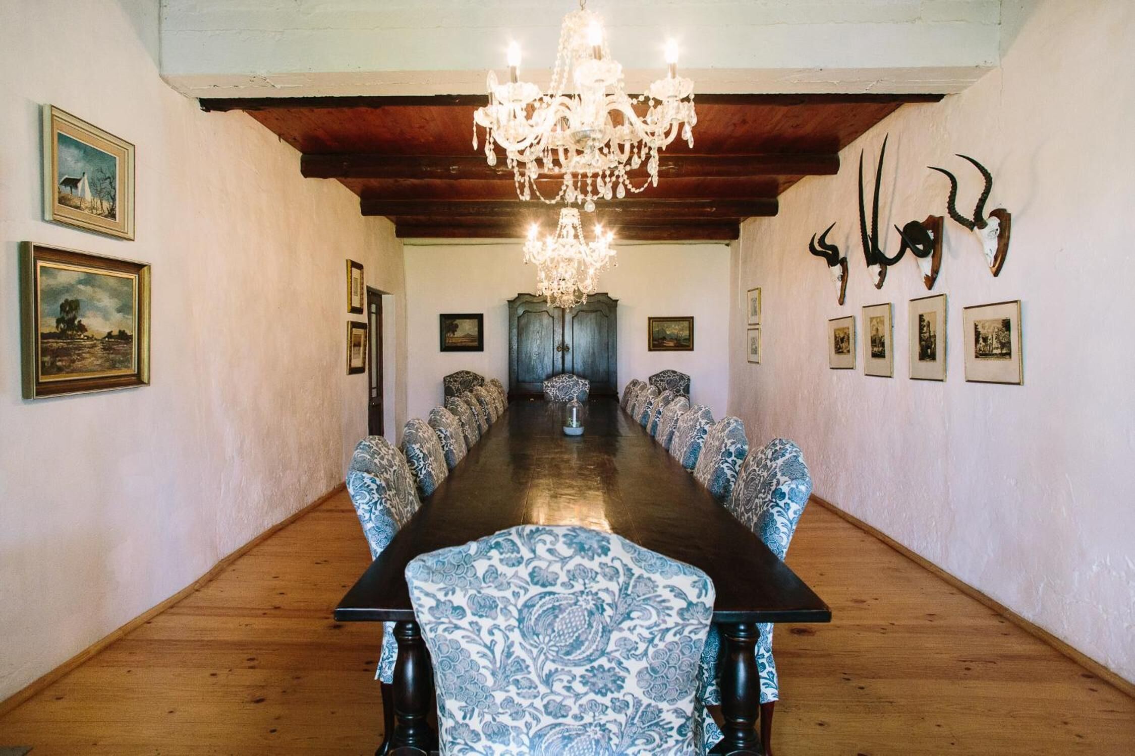 Une longue table à manger, des chaises à motifs, un lustre, des tableaux encadrés et un plafond en bois.