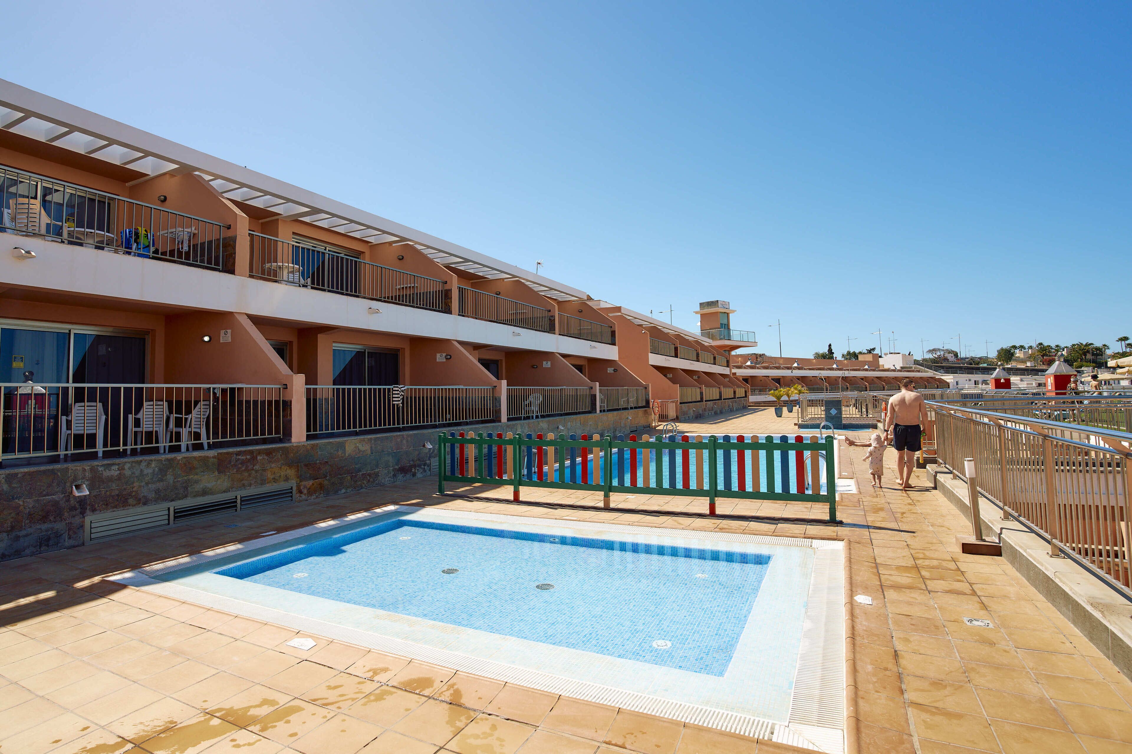 Zona de piscina con una piscina azul cristalina, una terraza con sol y barandilla, y un edificio con varios balcones.