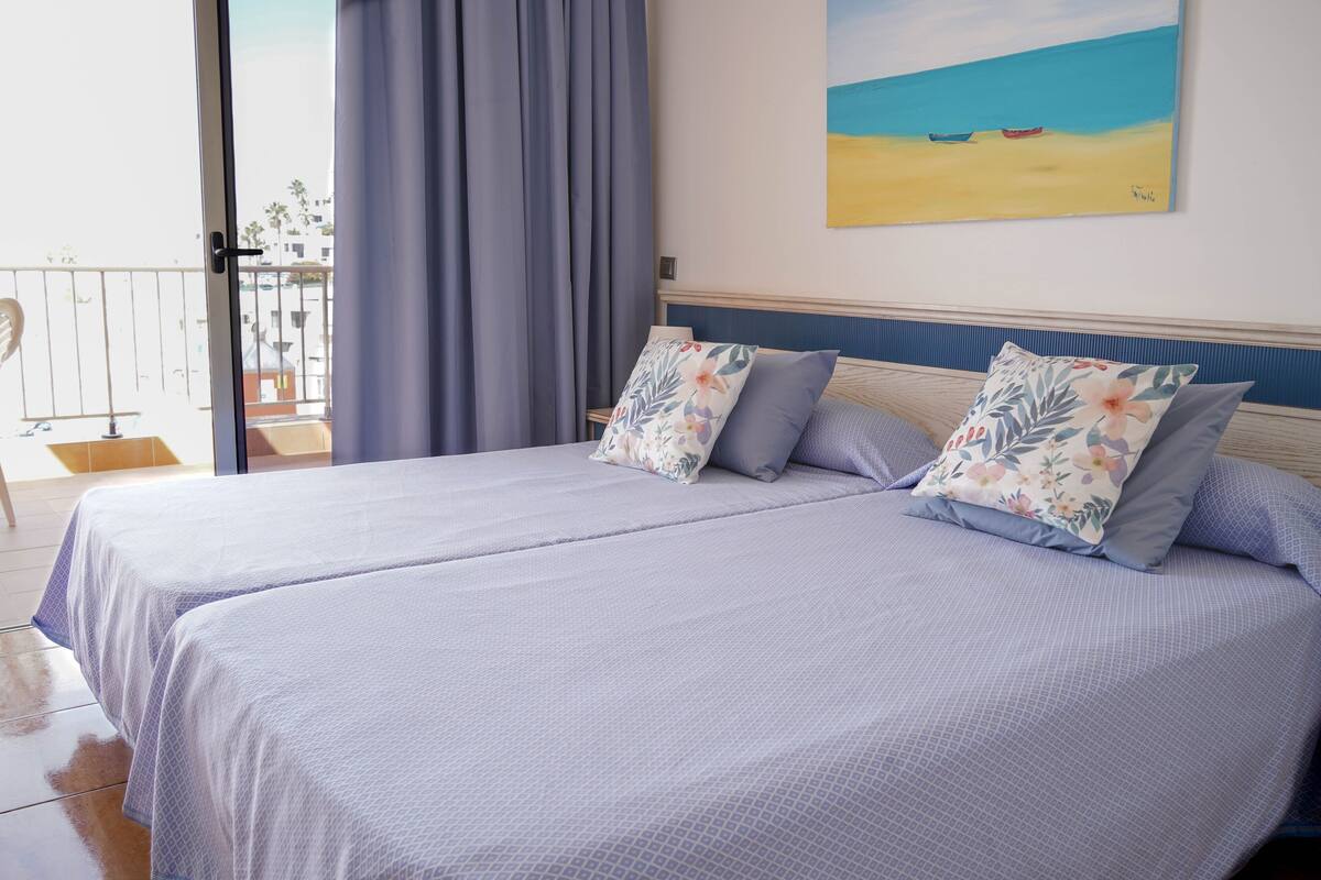 Un lit double agrémenté d’oreillers à motifs floraux, une vue sur la plage depuis un balcon et un tableau représentant des bateaux sur le sable.