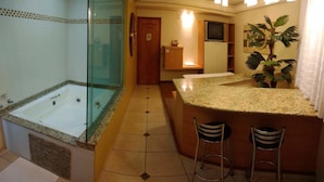 A bathroom with a glass-enclosed jacuzzi, a vanity with a sink, and a small bar area with stools. - Hotel Luni (São Paulo)